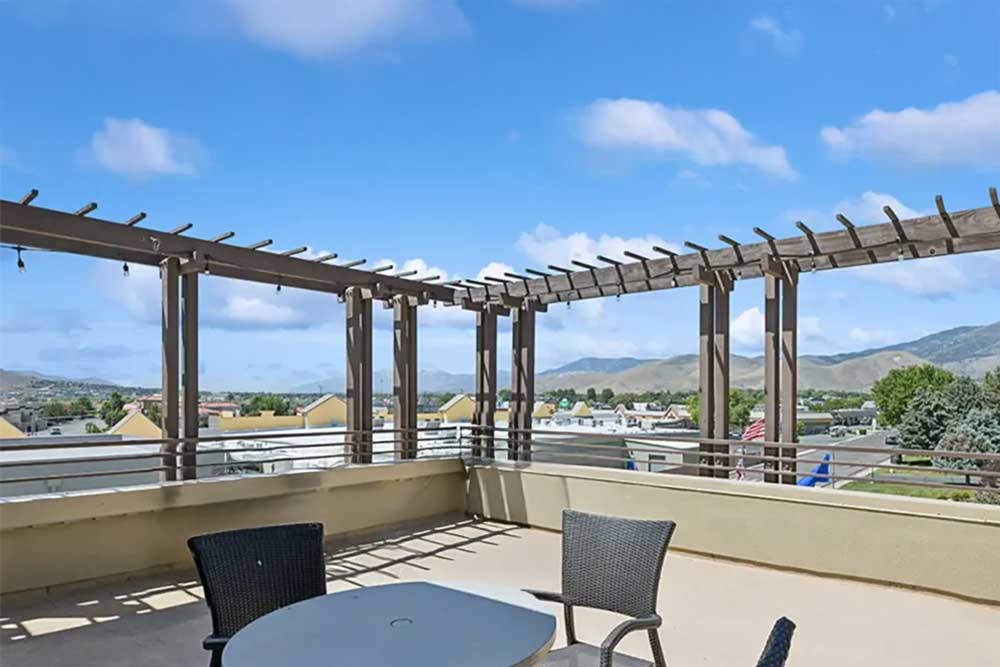 A rooftop patio with a table and chairs, featuring a wooden pergola overhead. The view overlooks rooftops, trees, and distant mountains under a partly cloudy blue sky.