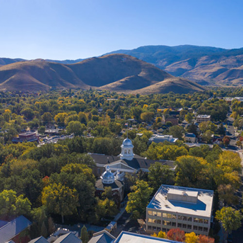 Aerial view of a small city with tree-lined streets, historic domed building, and surrounding mountains under a clear blue sky.