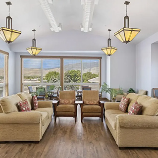Bright living room with two beige sofas, two armchairs, wooden coffee tables, and large windows with a scenic outdoor view. Four modern pendant lights hang from the ceiling, and there are plants and decorative pillows.
