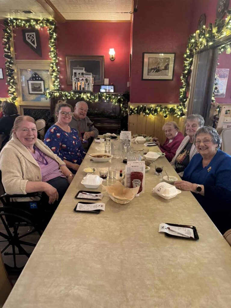 Six smiling seniors sitting around a rectangular restaurant table with bowls and drinks, surrounded by festive garlands and warm decor. Everyone appears to be enjoying a friendly meal together.