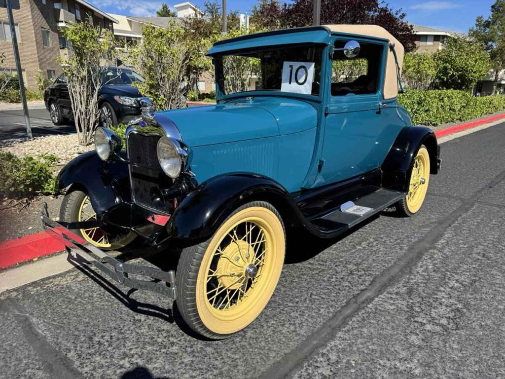 A vintage blue and black coupe with yellow wheels and a tan soft top is parked on a street, displaying the number 10 in the windshield. Trees and apartment buildings are visible in the background.