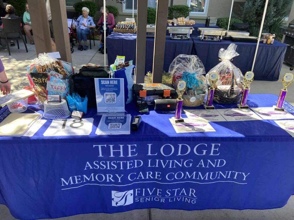 A blue tablecloth reads “The Lodge Assisted Living and Memory Care Community.” The table displays trophies, raffle baskets, treats, and flyers, with elderly people and food in the background at an outdoor event.