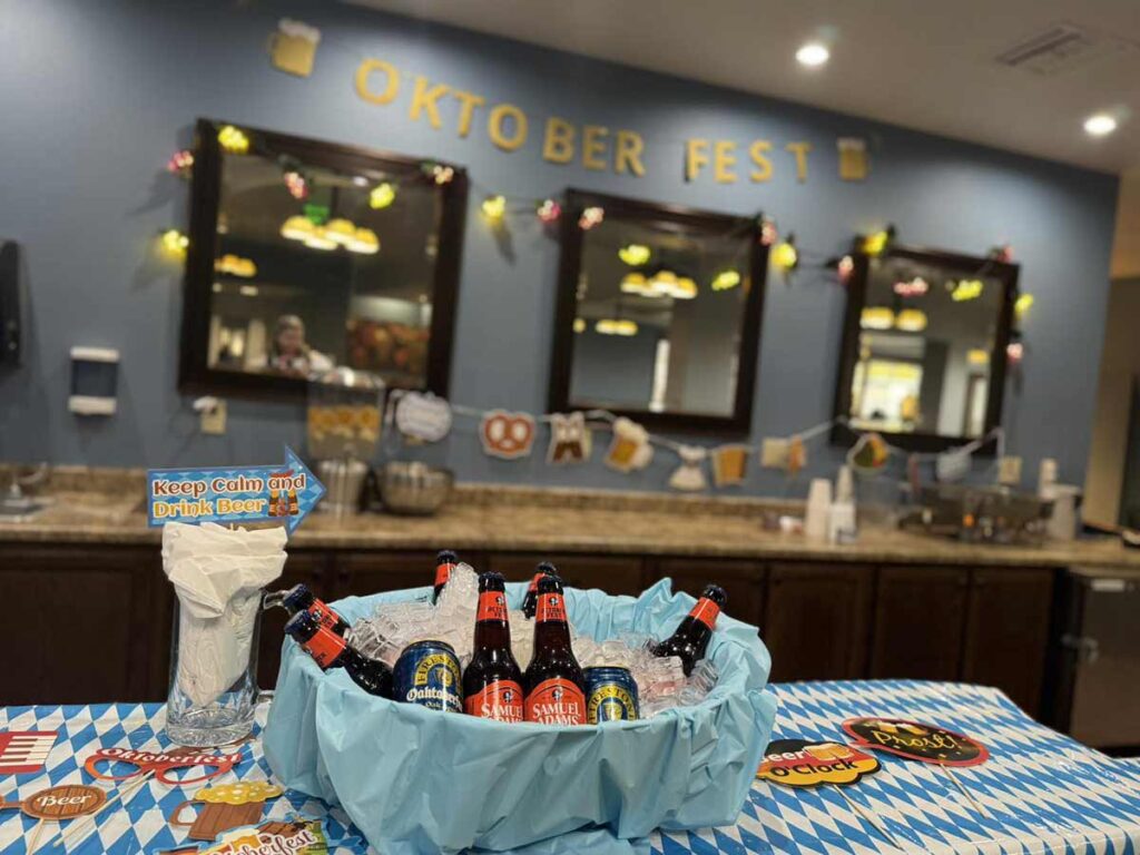 A table decorated for Oktoberfest with ice-filled bucket of bottled beers, a "Keep Calm and Drink Beer" sign, and festive blue-and-white cloth. The background has "Oktoberfest" banner and mirrors on the wall.