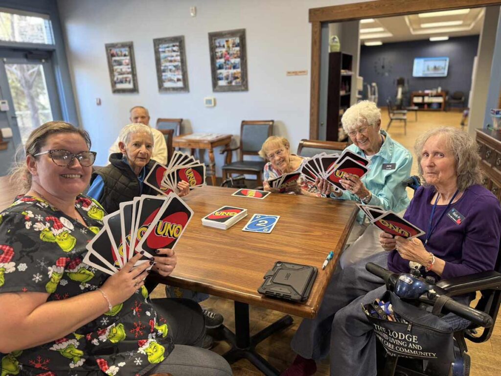 Six elderly people sit around a wooden table playing UNO with oversized cards, smiling and enjoying the game in a bright, cozy room at a senior living community.