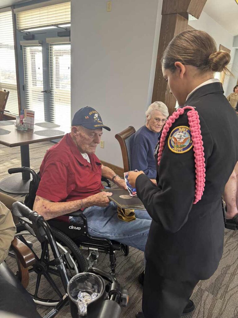 A young woman in uniform presents an award or ribbon to an elderly man in a wheelchair wearing a navy cap, while another elderly person sits nearby in a dining area.