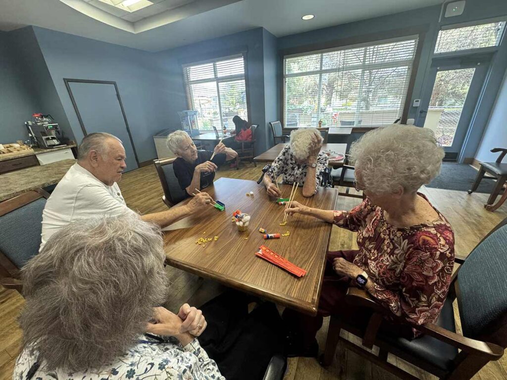 Five elderly people sit around a wooden table in a bright room, using chopsticks to pick up colorful plastic objects as part of an activity. Sunlight streams through large windows in the background.