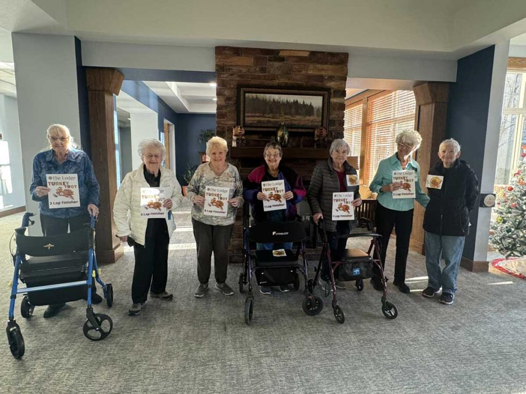 Seven elderly women stand indoors in a line, some using walkers, each holding a Thanksgiving-themed sign. They are smiling and the room is decorated warmly with a fireplace and windows in the background.
