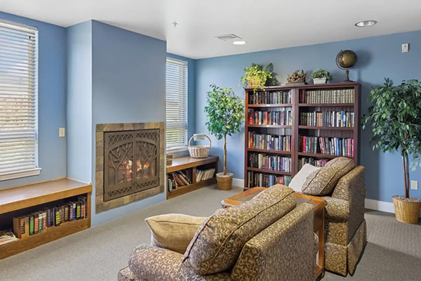 A cozy reading room with two patterned armchairs facing a fireplace, blue walls, large windows, potted plants, built-in shelves, and a tall bookcase filled with books and a globe.