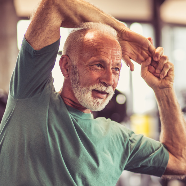 Older man with a white beard and mustache wearing a green t-shirt, smiling while stretching his arms overhead in a bright indoor setting.