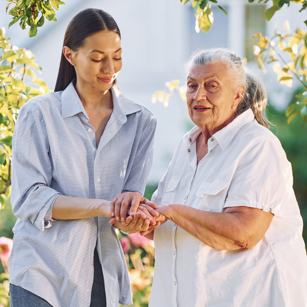 A young woman gently holds hands with an older woman outdoors in a garden, both wearing light-colored shirts and smiling warmly at each other.