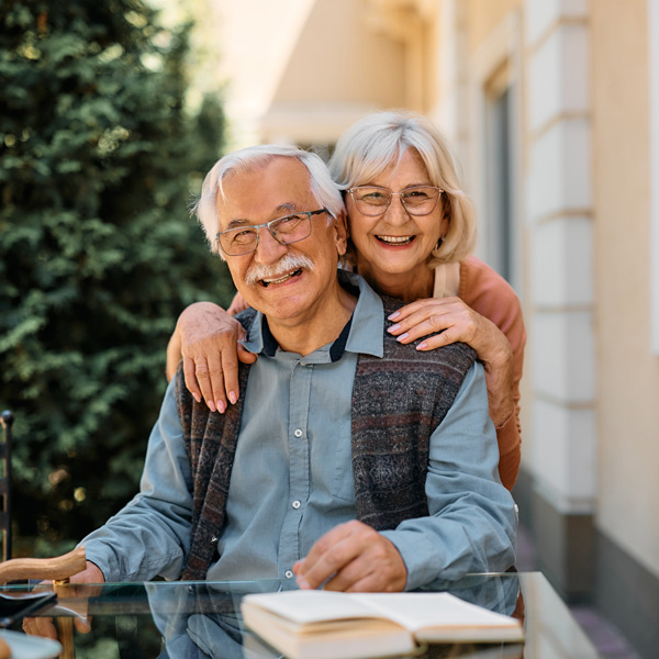 An elderly couple smiles warmly while sitting outdoors at a glass table with a book. The woman stands behind the man, hugging him from behind. They appear happy and relaxed, with trees and a building in the background.