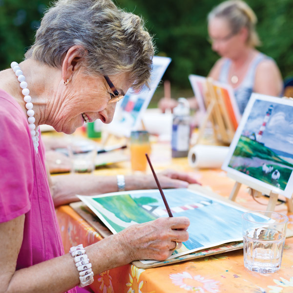 An older woman smiles while painting a watercolor landscape at an outdoor art class. She wears glasses, a pink shirt, and a beaded necklace. Another woman paints in the background. Paintings of lighthouses are visible.