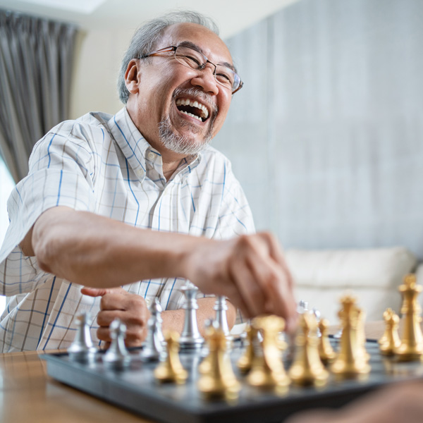 An elderly man wearing glasses and a plaid shirt smiles and laughs while playing chess, reaching out to move a piece on the chessboard in a bright living room.