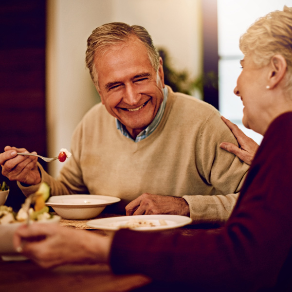 An older man and woman sit at a table, smiling and enjoying a meal together. The man holds a spoon with food, and they appear to be having a pleasant conversation in a cozy indoor setting.