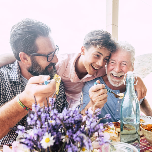 Three generations of men—grandfather, father, and son—are smiling and enjoying a meal together outdoors. A vase of purple flowers and a glass bottle sit on the table in the foreground.