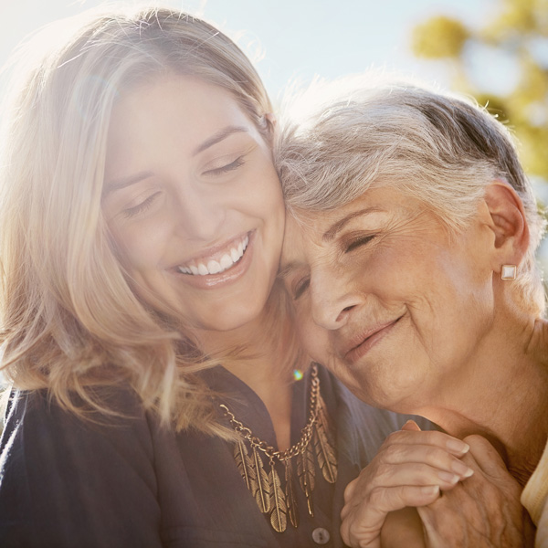 A smiling young woman and an older woman embrace outdoors in warm sunlight. The older woman rests her head on the younger woman’s shoulder, both appearing happy and content.