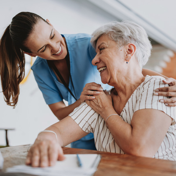 A smiling nurse in blue scrubs comforts and talks with an older woman with gray hair, who is seated at a table and laughing, creating a warm and supportive atmosphere.