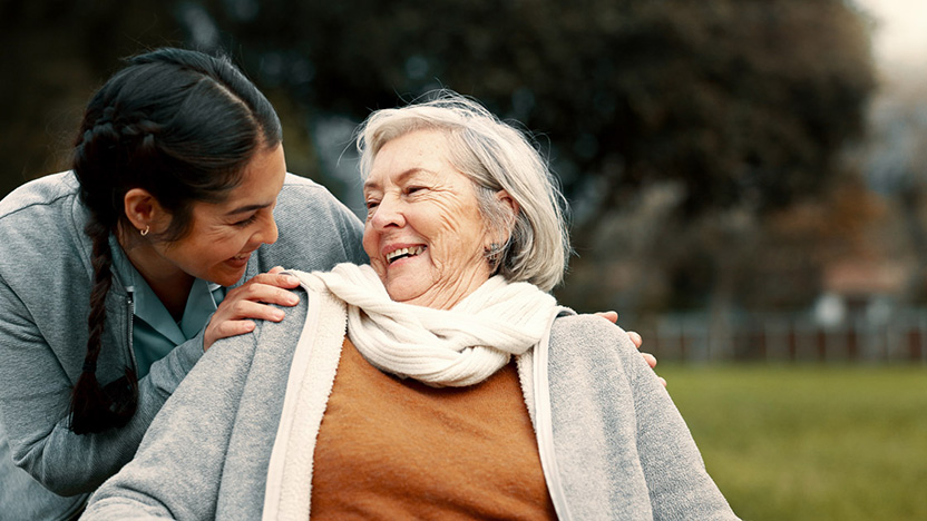 A young woman smiles warmly at an elderly woman in a wheelchair outdoors. Laughing in her white scarf and orange sweater, the elderly woman enjoys the moment—a heartwarming example of the benefits of memory care for loved ones.