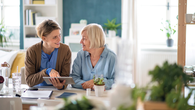 A younger woman, possibly a caregiver, and an older woman sit at a table together, smiling and talking. The younger woman holds a tablet, perhaps discussing the benefits of memory care as they enjoy a pleasant conversation in a bright kitchen.